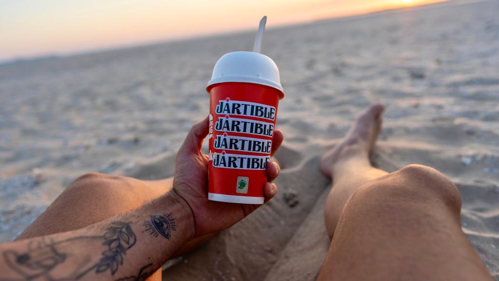 Vaso con logo de Jartible con la arena de la playa de fondo